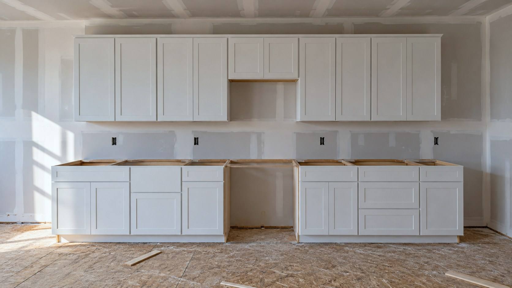 A new-house kitchen with plain white cabinets installed and no countertops yet, over a bare subfloor.
