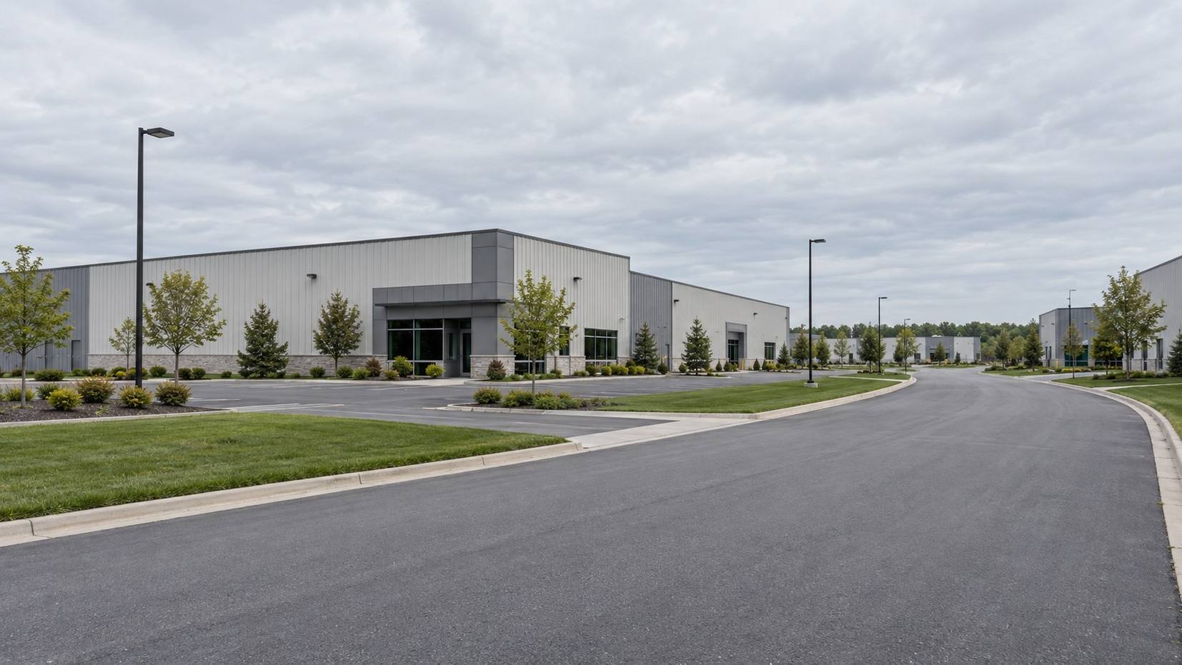 Road through an industrial park with plain manufacturing buildings, lawns and light poles on an overcast day