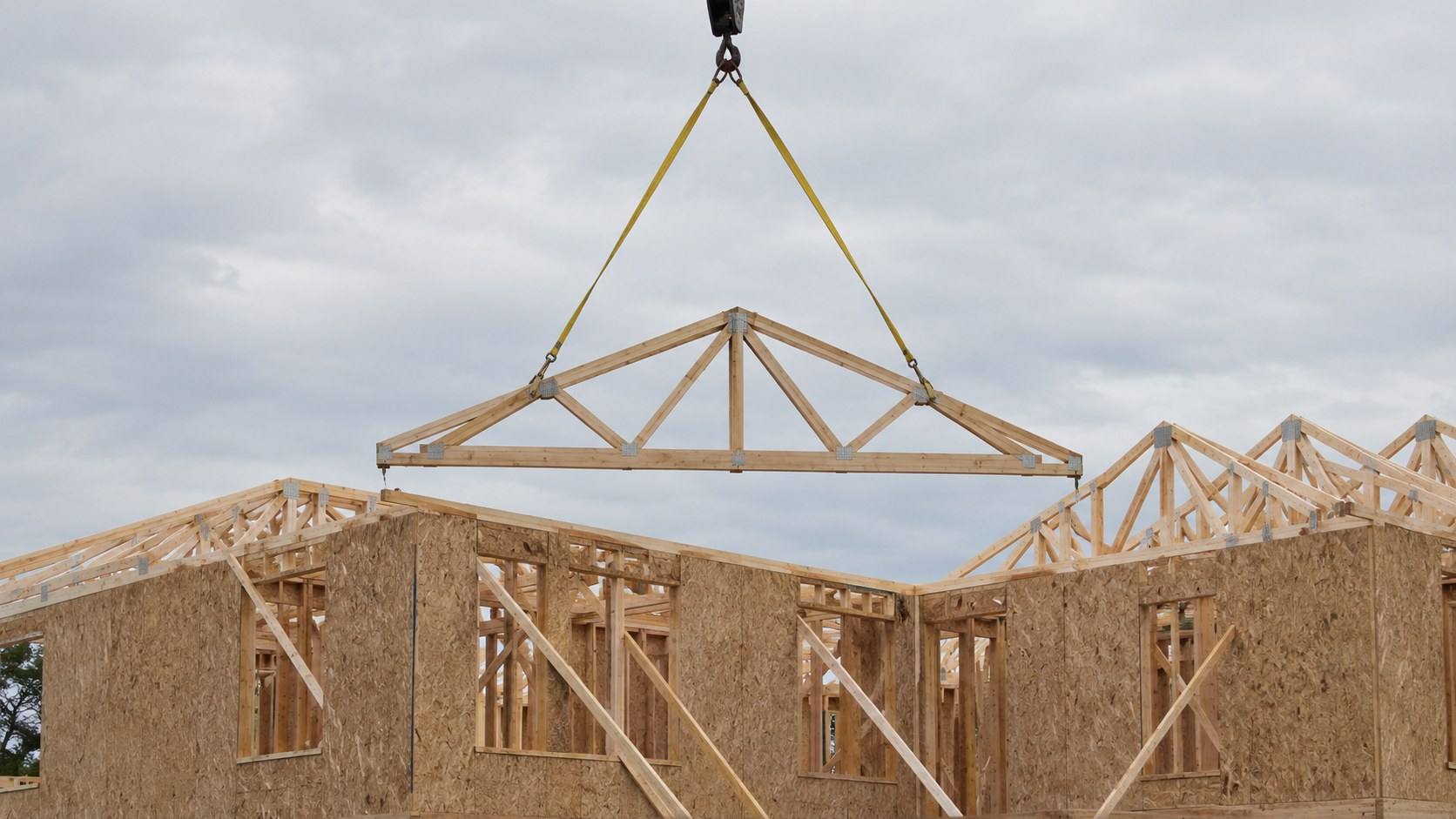 A wooden roof truss on a crane sling being lowered onto the walls of a framed house.